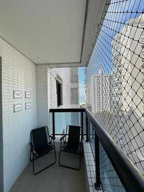 A balcony with two chairs, white tiled walls, and a view of buildings through a mesh screen. - SHB Paraíso em Pitangueiras  (Guarujá)