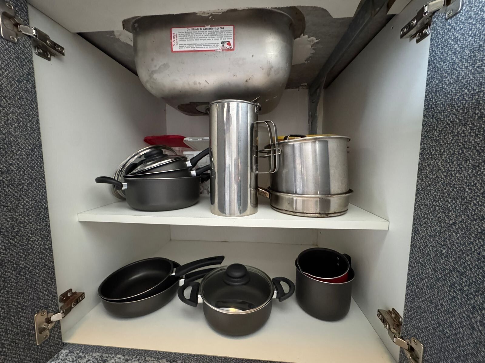A kitchen cabinet with cookware including pots, pans, and a kettle.
