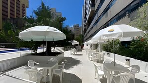 Mobiliario exterior con sillas y mesas de plástico blanco, sombrillas y una piscina al fondo.