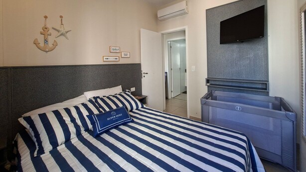 A hotel room with a bed, a flat-screen TV, an air conditioning unit, and a door leading to another room. - SHB Paraíso em Pitangueiras  (Guarujá)