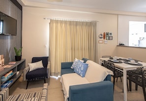 Una sala de estar con sofá, sillones, televisor, mesa de comedor con sillas y zona de cocina.