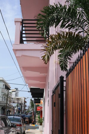 Un edificio rosa con un balcón, una entrada protegida por una reja y coches estacionados a lo largo de la calle.