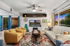 A modern living room with a ceiling fan, a flat-screen TV, a fireplace, and a large window with a view. - 1962 TramView Palm Springs - Poolside Fun, Spa, Fire Pit & Views (Palm Springs)
