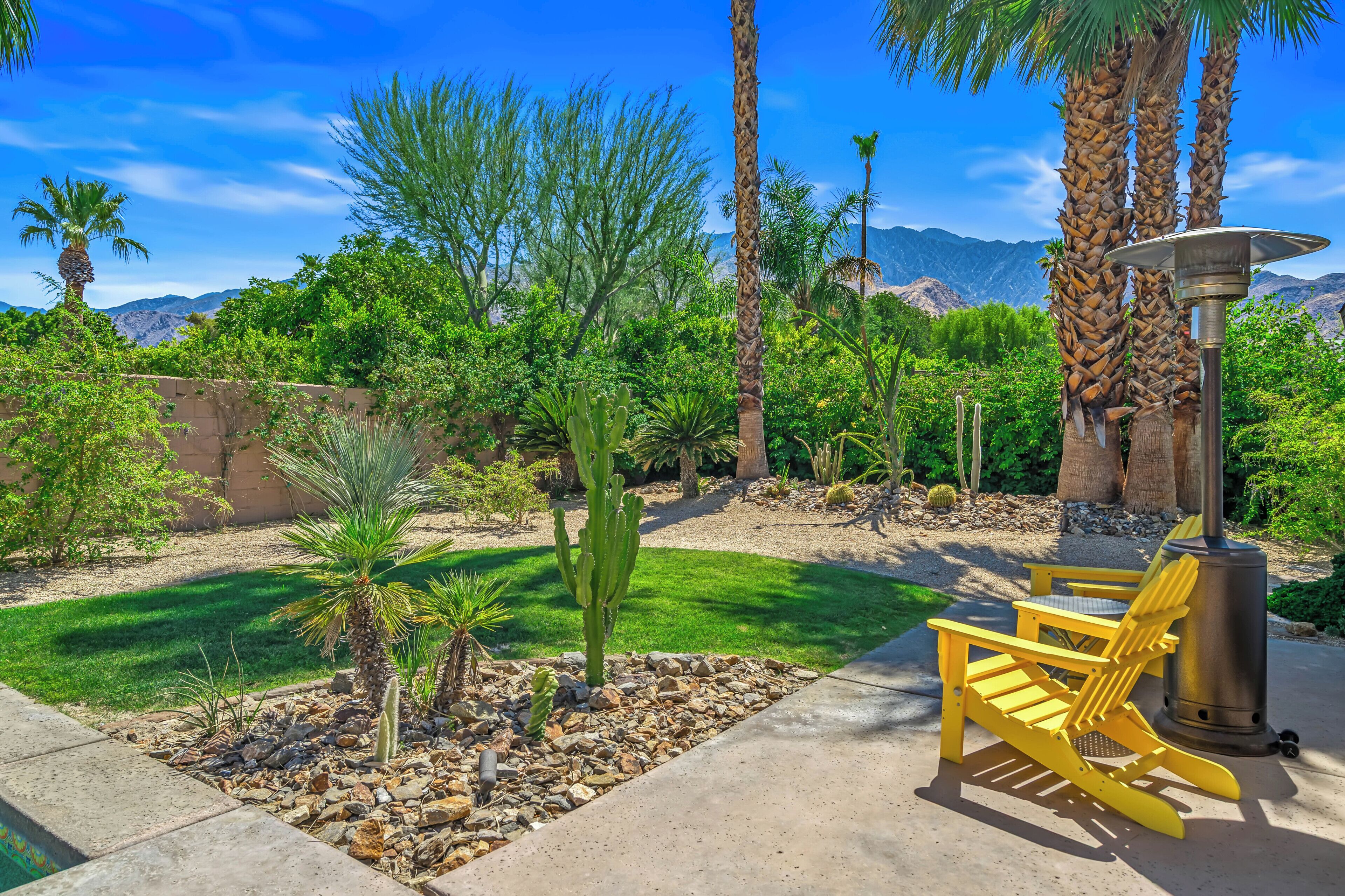 A patio area with a concrete floor, yellow chairs, a metal heater, and a variety of desert plants.