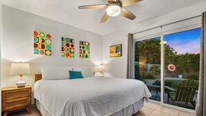 A bedroom with a large bed, two bedside tables, a ceiling fan, and artwork on the walls. - 1962 TramView Palm Springs - Poolside Fun, Spa, Fire Pit & Views (Palm Springs)