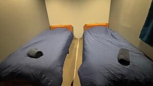 A narrow room with two single beds, each with a wooden headboard and a blue mattress cover.