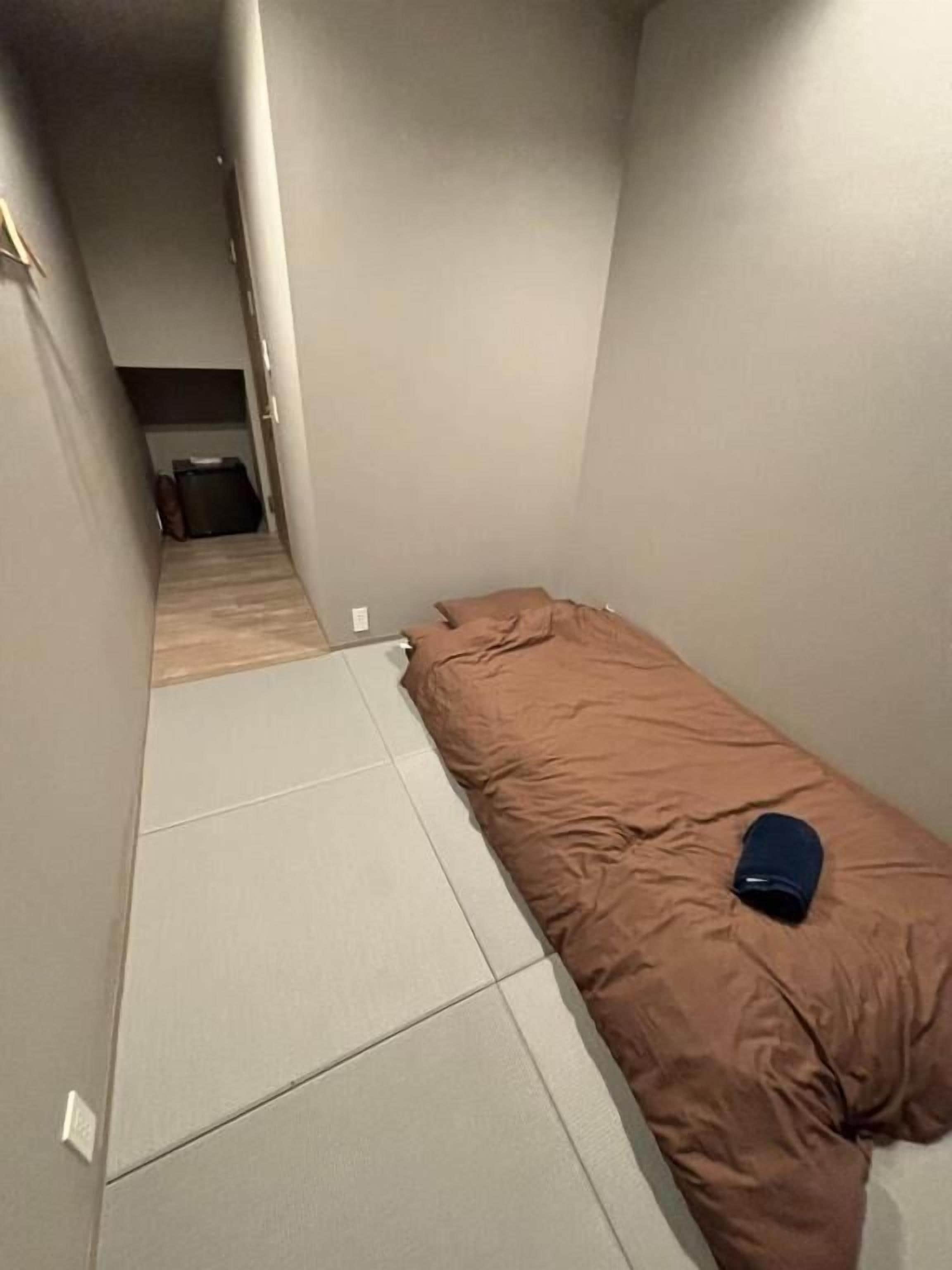 Un pasillo estrecho con una cama a la derecha, una maleta al fondo y una puerta al final.