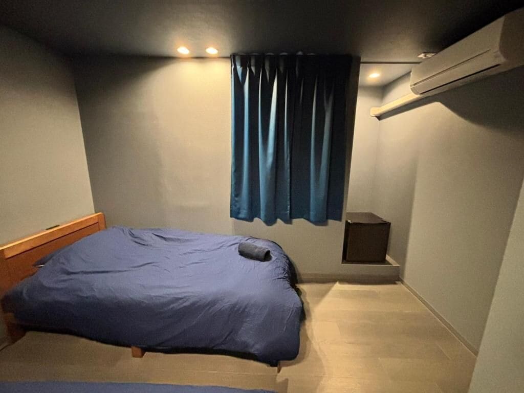 Una habitación pequeña con cama, una ventana con cortinas, un aire acondicionado de pared y un altavoz pequeño.