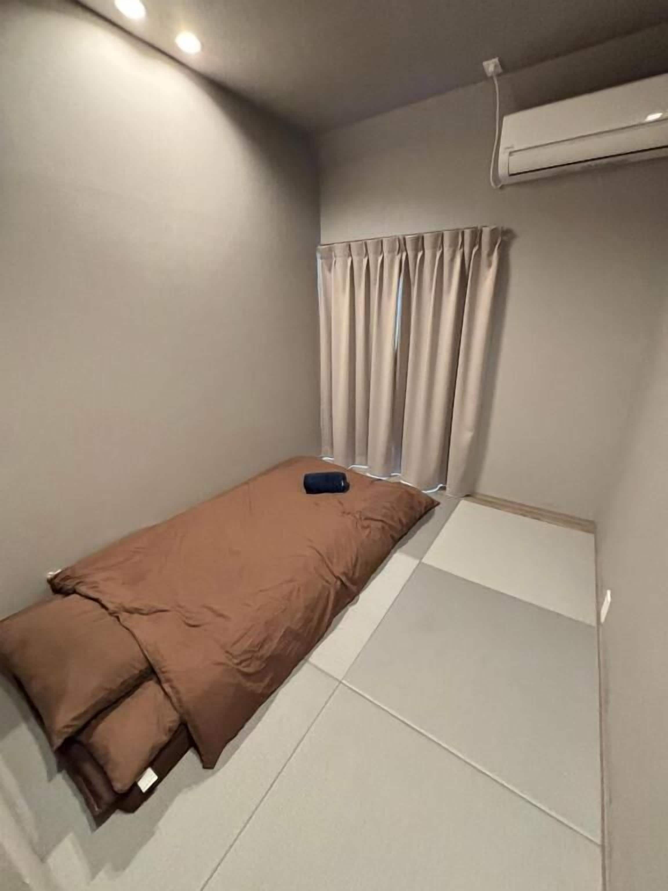 Une petite chambre avec un lit simple, une fenêtre avec des rideaux, un appareil de climatisation et un luminaire fixé au mur.