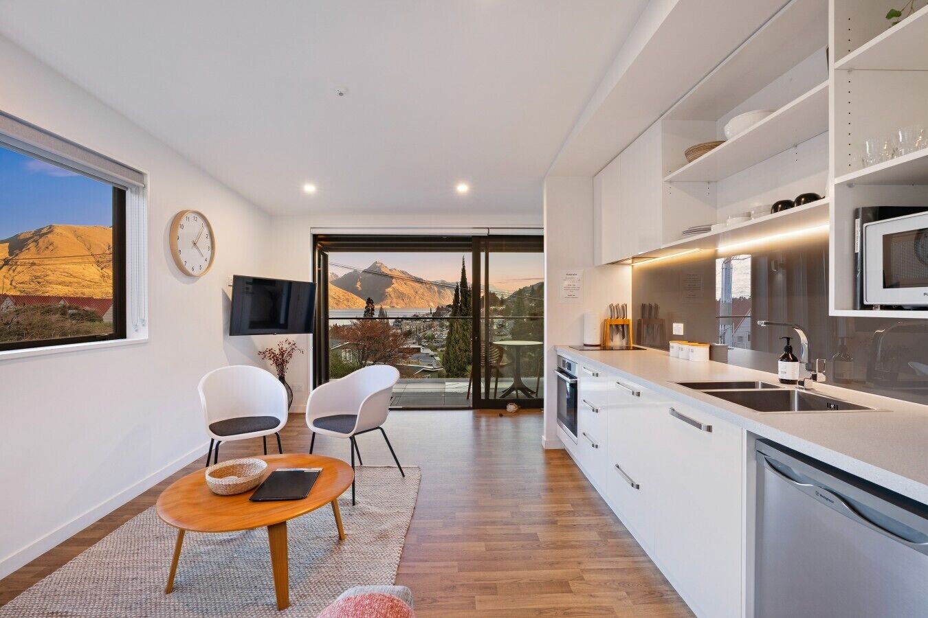 A modern kitchen with a large island, a dining area with chairs, a TV mounted on the wall, and a view of mountains through a glass door.