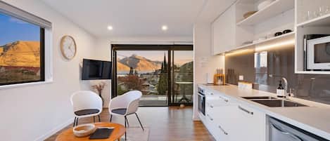 A modern kitchen with a large island, a dining area with chairs, a TV mounted on the wall, and a view of mountains through a glass door.