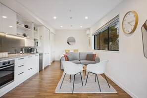 A modern kitchen with white cabinetry, a dining area with a grey sofa and white chairs, and a large window with a view. - Queenstown Quarters - Residence IV (Queenstown)