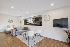 A modern living room with a sofa, chairs, a coffee table, a large window, a wall clock, and a flat-screen TV. - Queenstown Quarters - Residence IV (Queenstown)