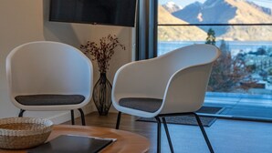 Quarto moderno com duas cadeiras brancas, uma mesa de madeira, um cesto de palha e uma grande janela com vista para as montanhas.