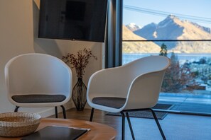Ein modernes Zimmer mit zwei weißen Stühlen, einem Holztisch, einem gewebten Korb und einem großen Fenster mit Blick auf die Berge.