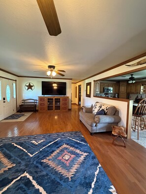 A spacious living room with a large TV, wooden floors, a ceiling fan, and a kitchen area visible through an open doorway. - The Cowboy Way a Charming farmhouse with stunning ranch views and HUGE GAME ROOM (Gallipolis)