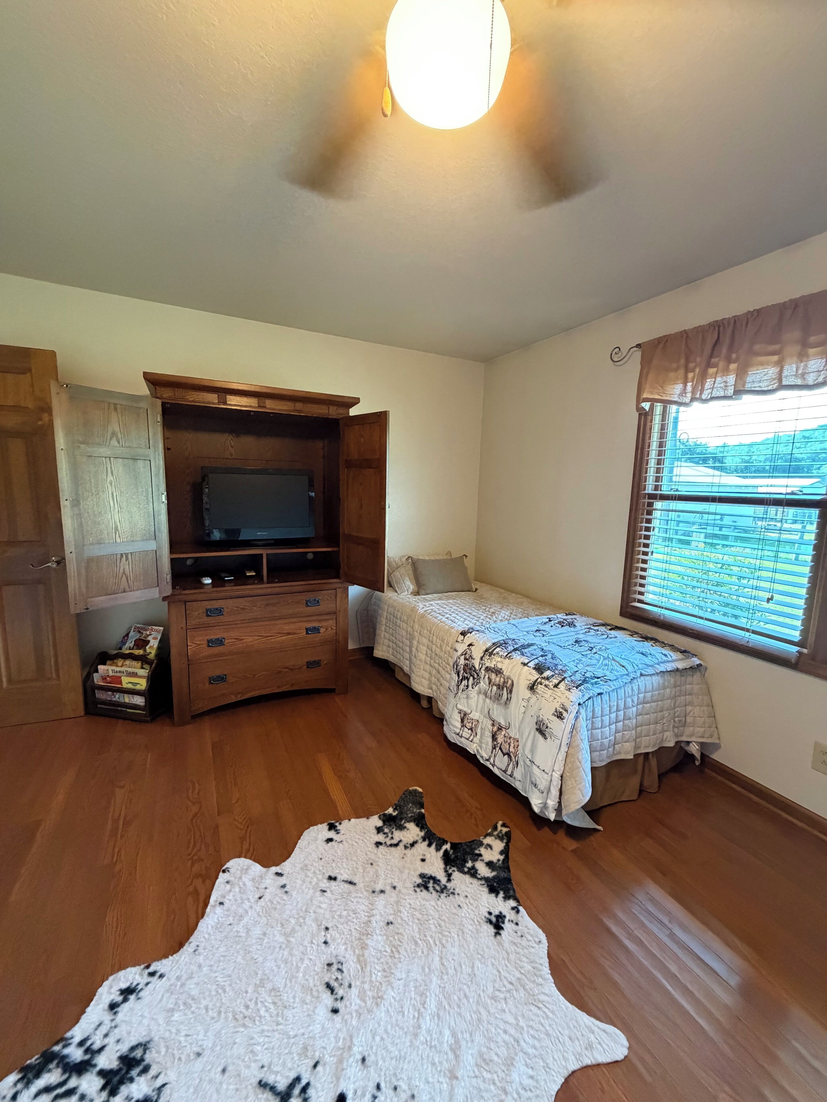 A bedroom with a wooden dresser, a bed with a patterned bedspread, a window with blinds, and a ceiling light.