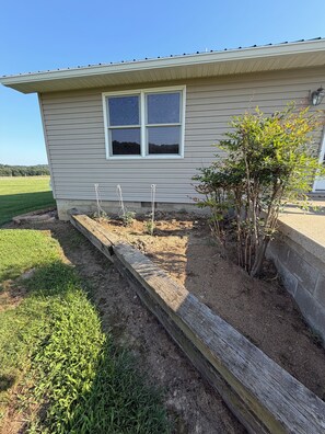 Een huis met een raam, een kleine tuin met planten en een houten rand, en een heldere lucht.