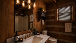 Ein Badezimmer in einer Holzhütte mit einem großen Spiegel, einem Waschbecken mit Marmorplatte, einer Toilette und einem Fenster mit Jalousien.