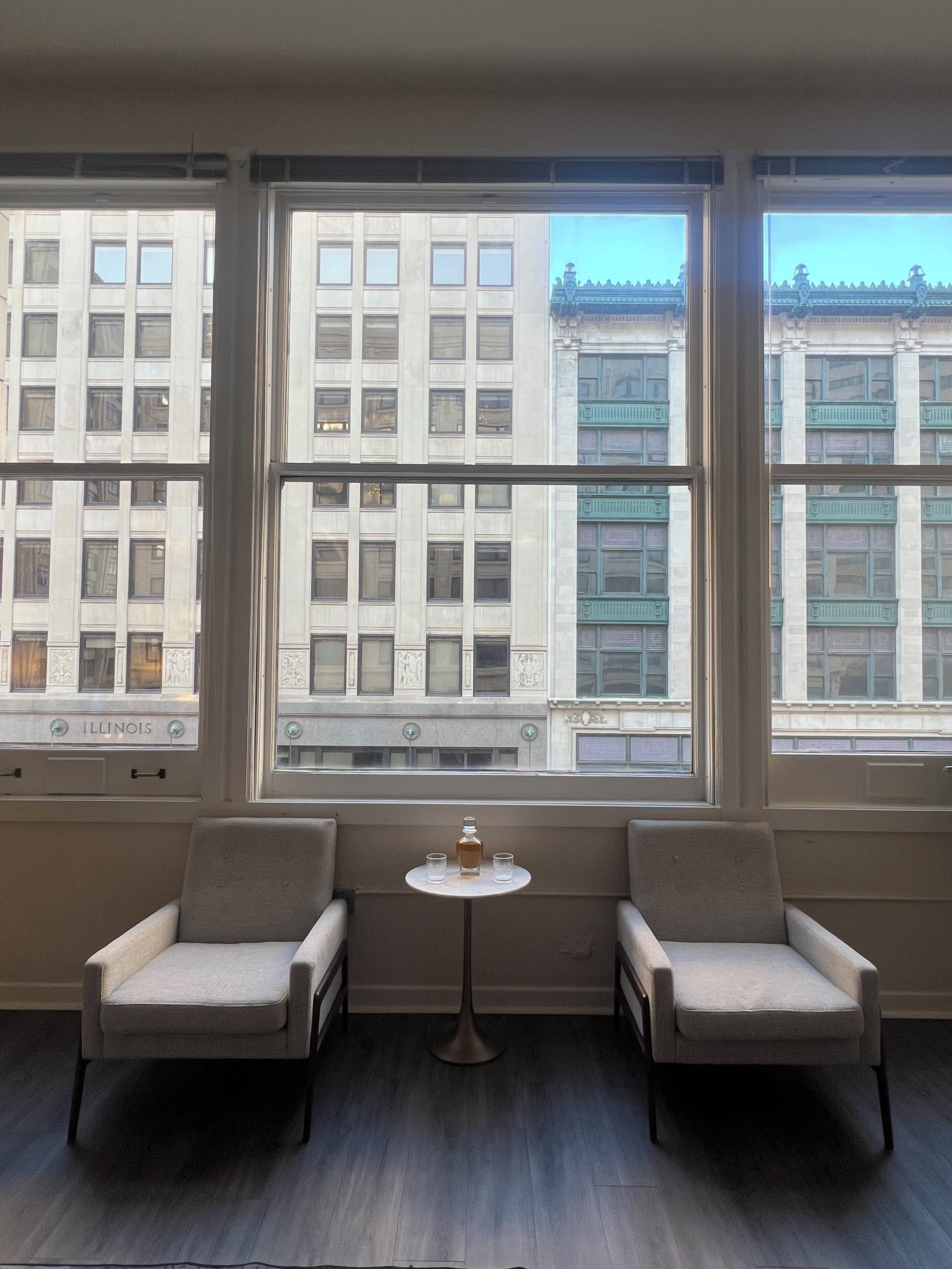 Una habitación con dos sillones y una mesita, ventanas amplias con vista a un edificio que muestra la palabra 'ILLINOIS'.