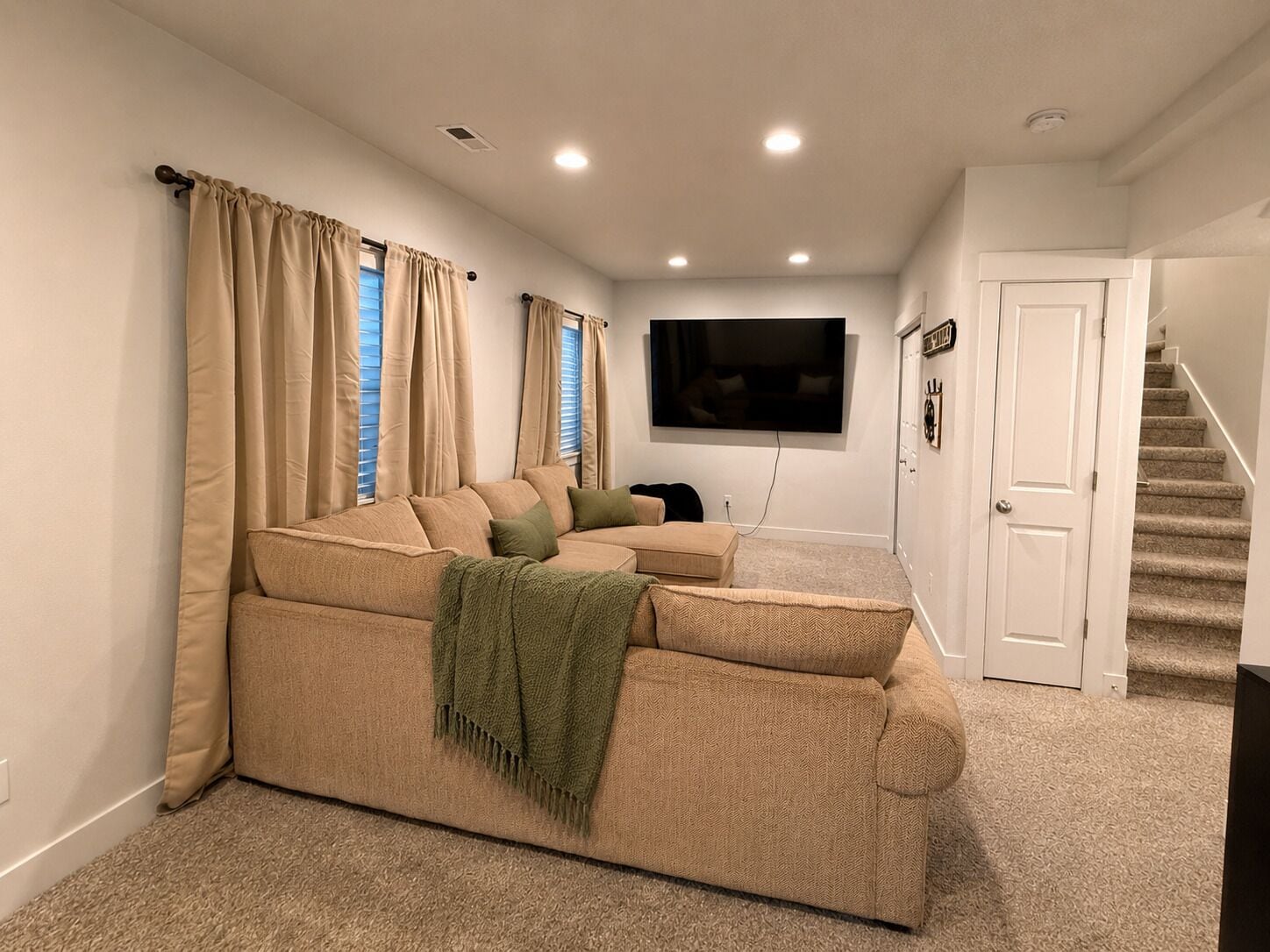 A living room with a beige sectional sofa, a flat-screen TV mounted on the wall, and a staircase leading to an upper level.