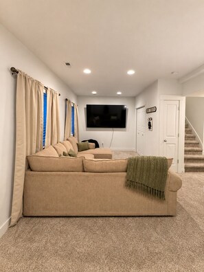 A living room with a beige sofa, a flat-screen TV, beige curtains, and a staircase. - The Point Retreat, Theater, Firepit, Near I-15 (Bluffdale)