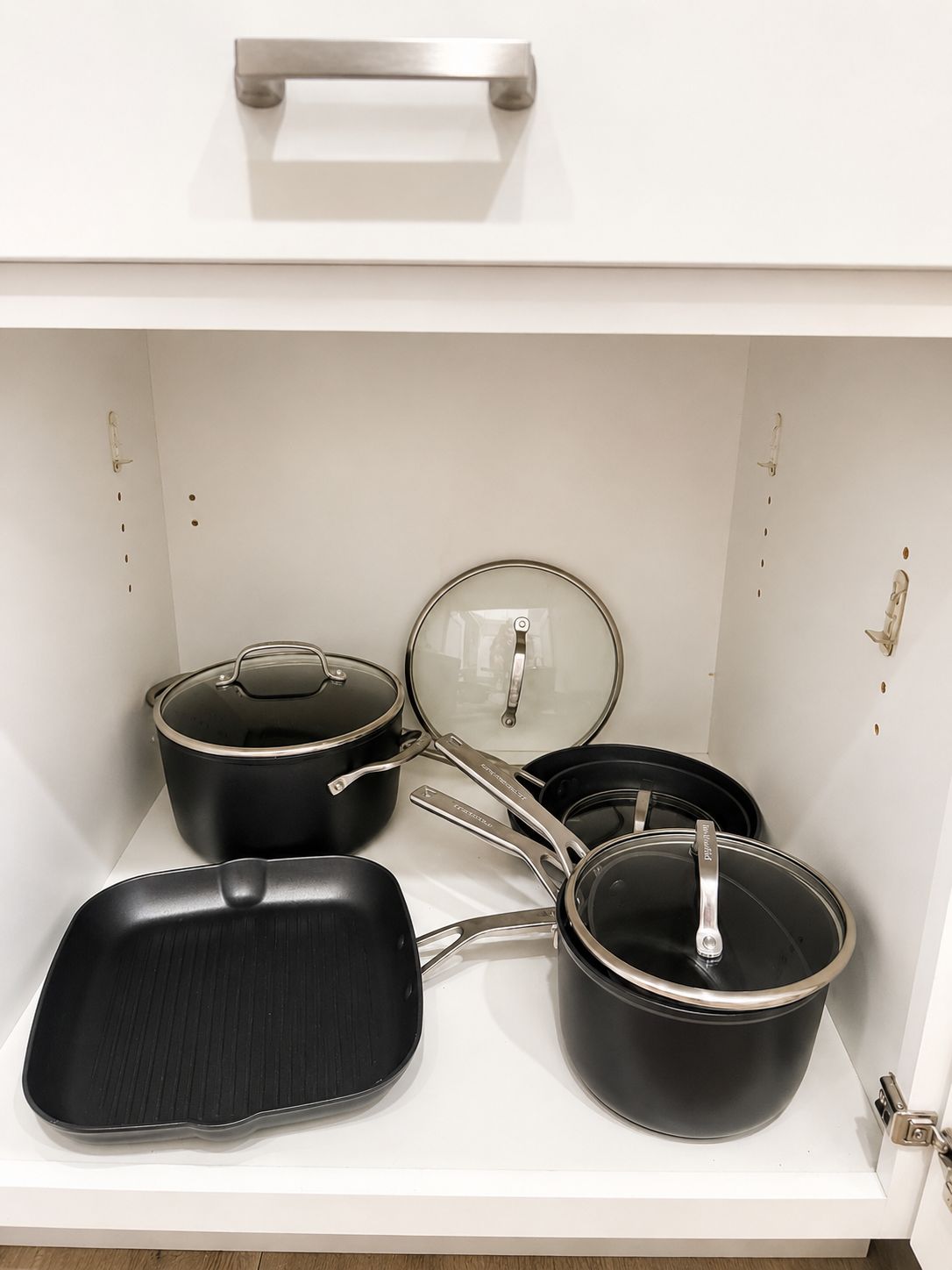 Armário de cozinha com puxador, contendo diversos utensílios de cozinha, incluindo panelas, frigideiras e uma assadeira.