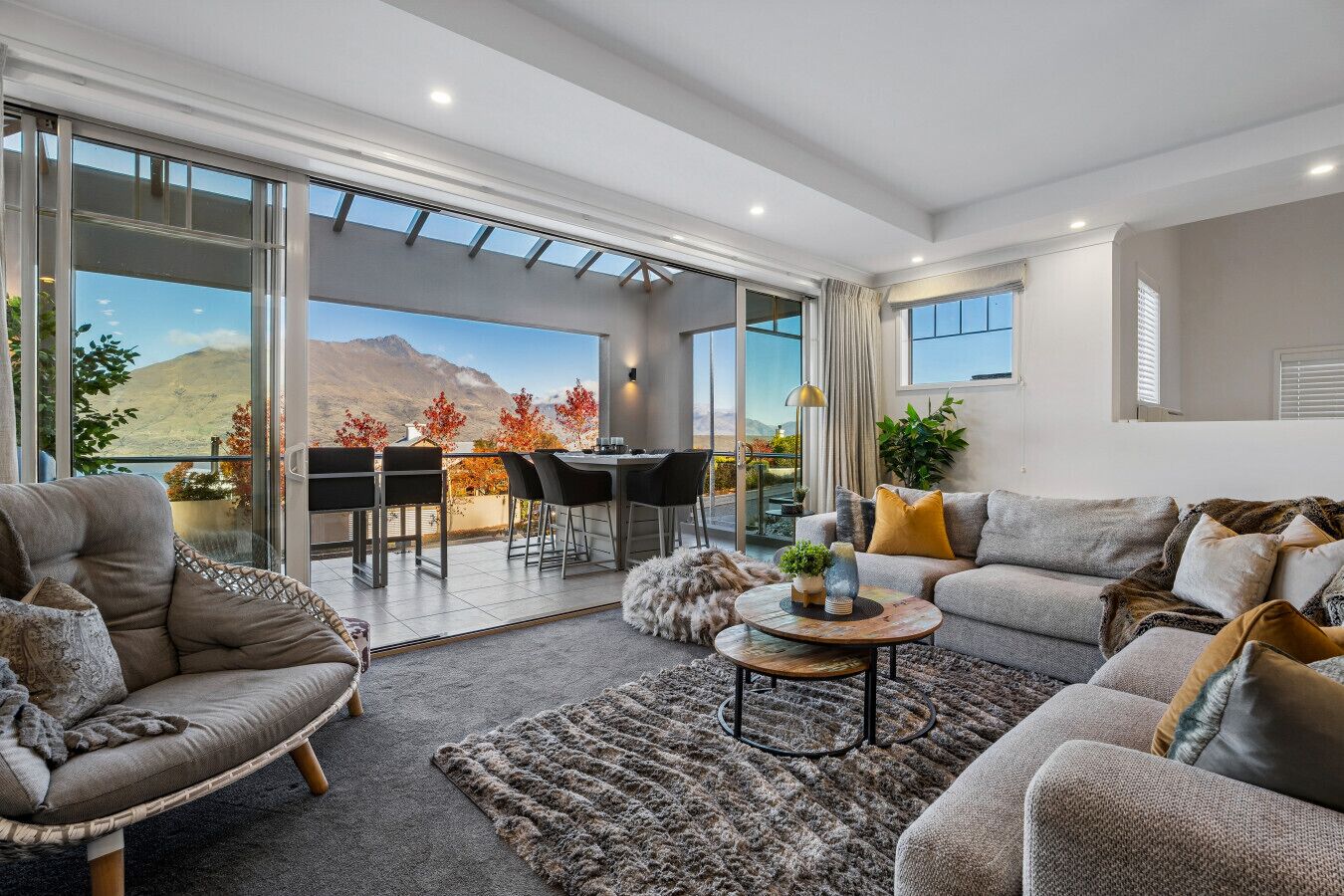 A modern living room with a sofa, armchair, and coffee table, a balcony with a dining area, and a mountain view.