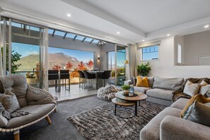A modern living room with a sofa, armchair, and coffee table, a balcony with a dining area, and a mountain view. - Remarkable 5-Bedroom on the Hill (Queenstown)