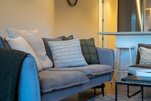 Ein Wohnzimmer mit einer Couch, einem Couchtisch und einem Sessel. Im Hintergrund ist eine Küchenzeile zu sehen.