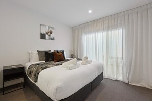 Ein modernes Hotelzimmer mit einem großen Bett, einem Nachttisch, einem Beistelltisch, einem Bild an der Wand und durchsichtigen Vorhängen am Fenster.