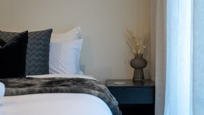 Een netjes opgemaakt bed met een donkere deken en kussens, een nachtkastje met een vaas gedroogde planten, en uitzicht op een raam met dunne gordijnen.