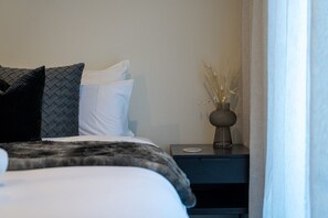 A neatly made bed with a dark blanket and pillows, a bedside table with a vase of dried plants, and a view of a window with sheer curtains. - Remarkable New Home on the Hill (Queenstown)