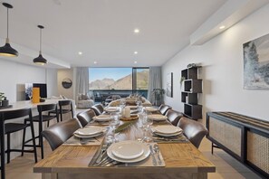 A dining area with a long table set for six, surrounded by leather chairs, with a view of mountains through large windows. - Remarkable New Home on the Hill (Queenstown)