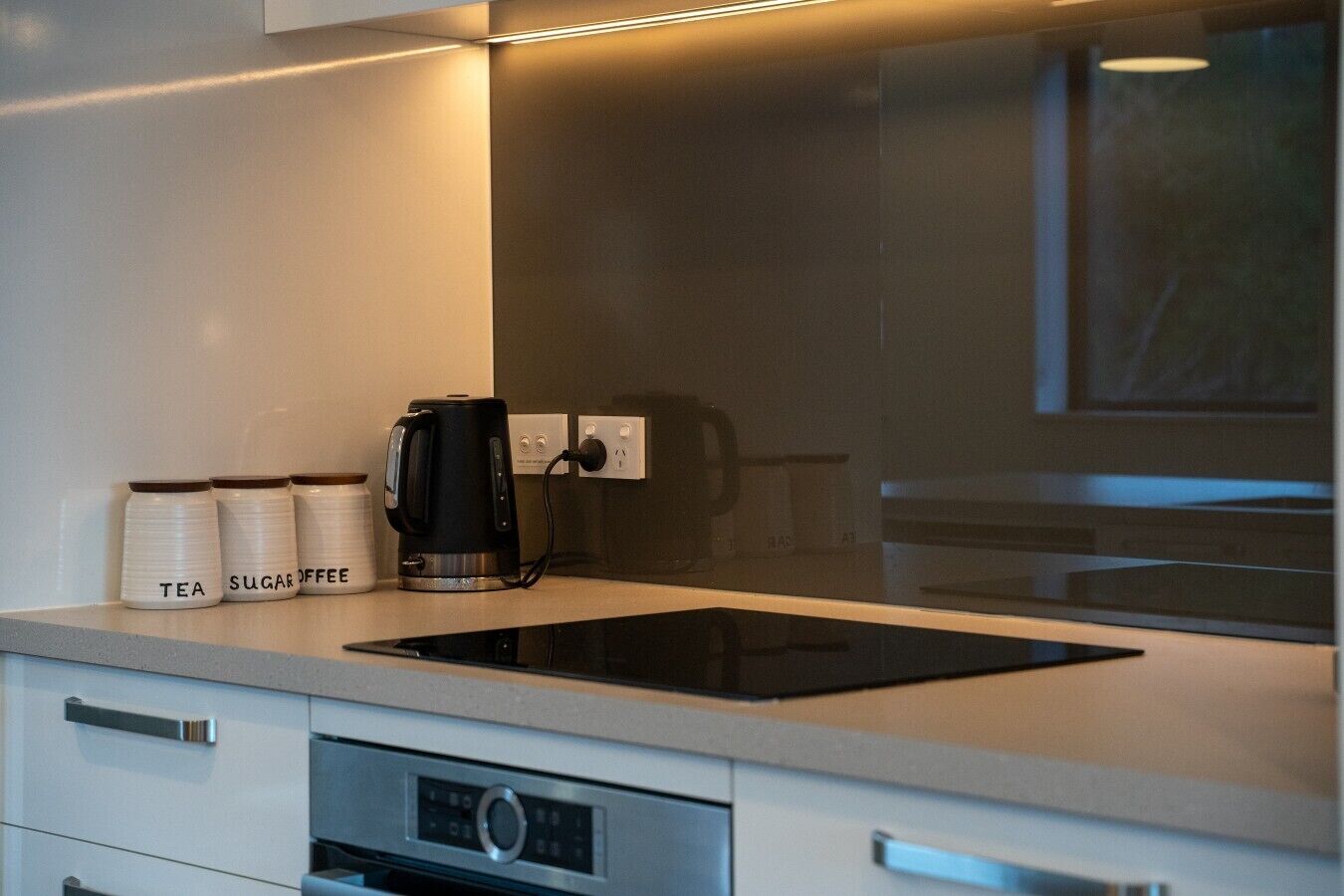 Una cocina con placa de inducción negra, cafetera y recipientes etiquetados para té y azúcar.