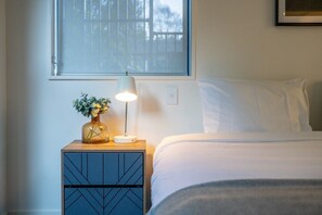 A bedroom with a bed, a nightstand with a lamp, a vase with flowers, and a window with blinds. - Queenstown Quarters - Residence III (Queenstown)