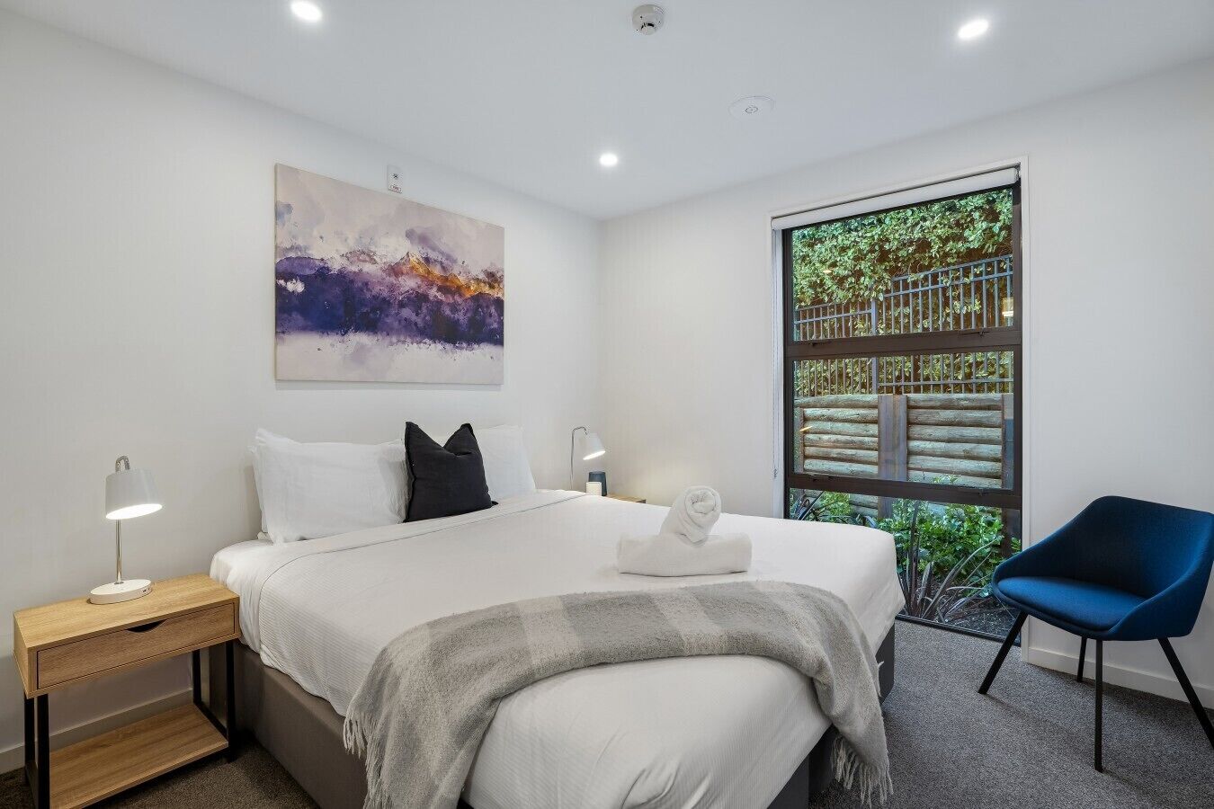 Une chambre d’hôtel moderne avec un grand lit, une table de chevet, une chaise, une fenêtre donnant sur un espace verdoyant et un tableau au mur.