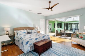 A bedroom with a large bed, a nightstand, a lamp, a ceiling fan, and a view of a green house through a sliding glass door. - Min from Naples in Beautiful Brendan Cove w/Private Pool and Covered Courtyard (Bonita Springs)