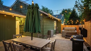 Un patio al aire libre con mesa y sillas de madera, una parrilla y luces colgantes.
