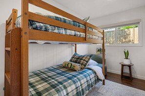 A bunk bed with a plaid bedspread and pillows, a small bedside table with a plant, and a window with a view of the outside.