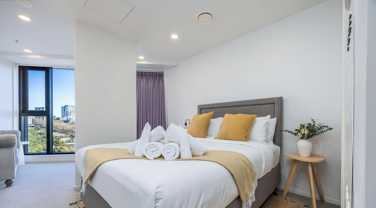 A hotel room with a bed, a window with a view, a bedside table with a plant, and a chair.