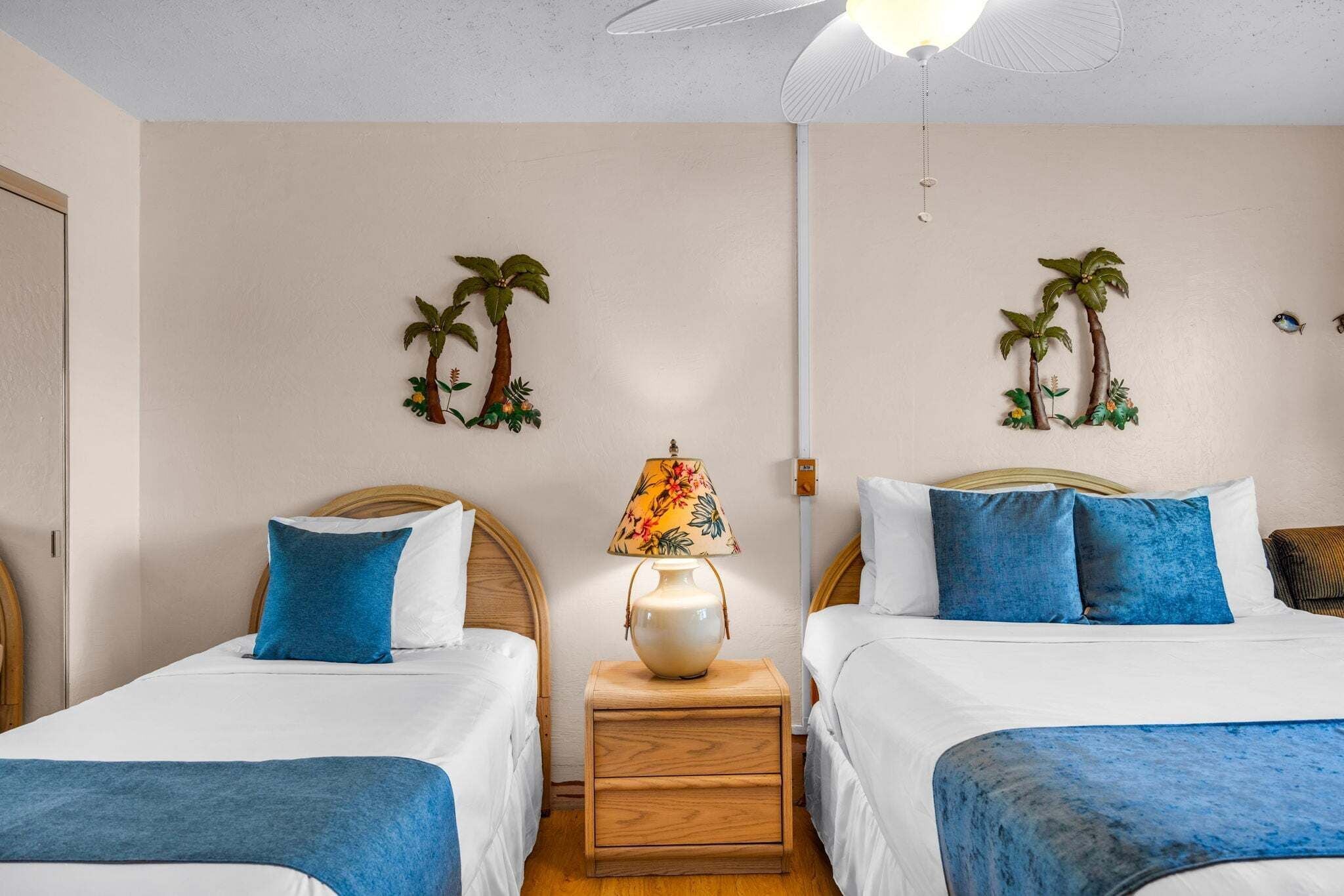 A hotel room with two beds, a nightstand with a lamp, and wall decorations of palm trees.