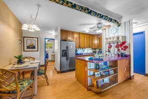 A modern kitchen with wooden cabinets, a central island, and a dining area with chairs. - Charming Tropical 2BR Condo in Kihei (Kihei)