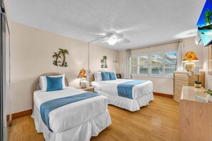 Ein Hotelzimmer mit zwei Betten, einem Deckenventilator, einem Fenster mit Jalousien, einem Schreibtisch mit Lampe und einem an der Wand befestigten Fernseher.