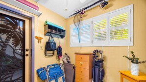 A room with a door, a window with blinds, a wooden cabinet, a blue chair, a golf bag, and a small table with a plant. - Charming Tropical 2BR Condo in Kihei (Kihei)