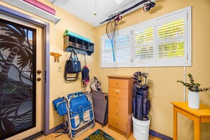 A room with a door, a window with blinds, a wooden cabinet, a blue chair, a golf bag, and a small table with a plant. - Charming Tropical 2BR Condo in Kihei (Kihei)