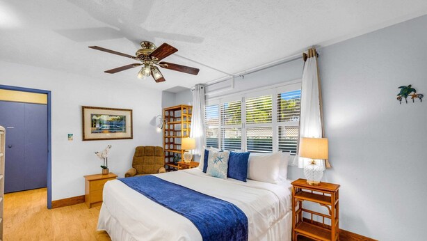 A bedroom with a bed, a ceiling fan, a window with blinds, a nightstand, a chair, and a wall-mounted shelf. - Charming Tropical 2BR Condo in Kihei (Kihei)