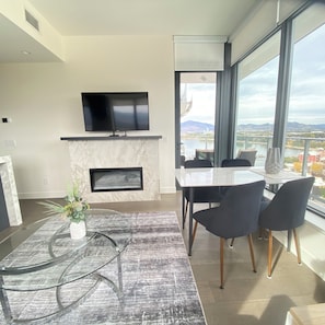 A modern living room with a flat-screen TV, a fireplace, a dining area with chairs, and a large window offering a view of a cityscape. - Aqua Waterfront Resort (Kelowna)