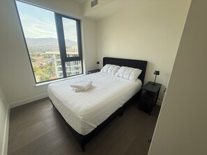 A bedroom with a large window, a bed with white bedding, a nightstand, and a lamp. - Aqua Waterfront Resort (Kelowna)