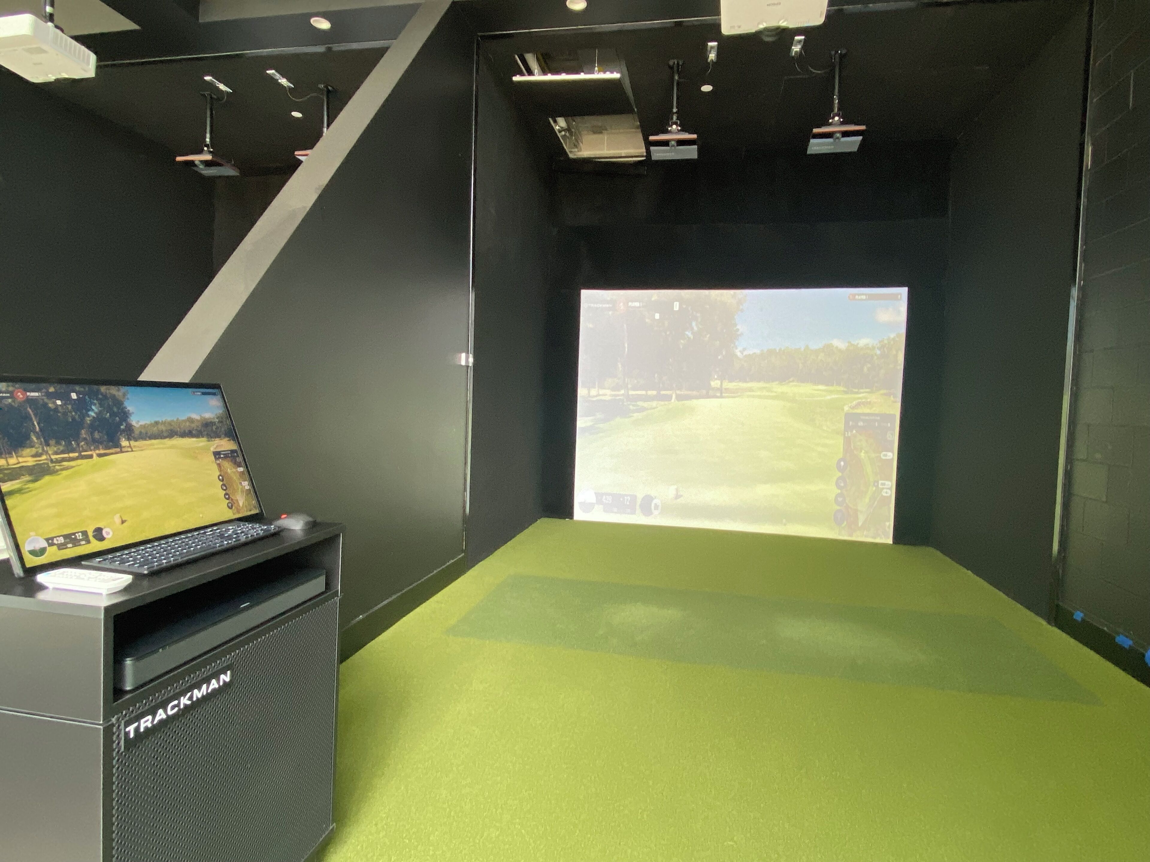 A room with a green carpet, a mounted screen displaying a golf game, and a speaker labeled 'TRACKMAN'.